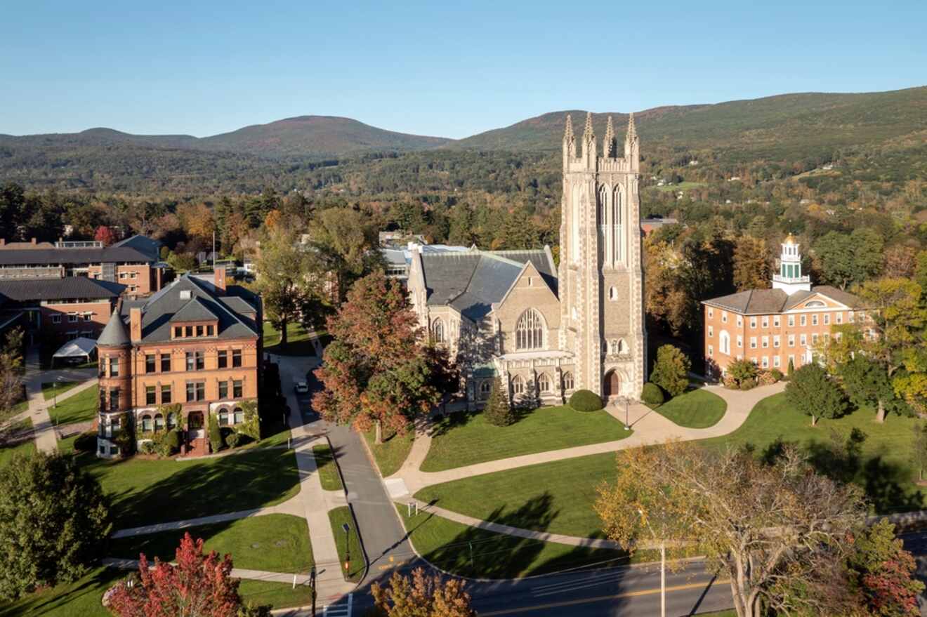 Aerial view of a college campus with a large stone church, surrounding buildings, walkways, and green spaces, set against a backdrop of rolling hills and trees.