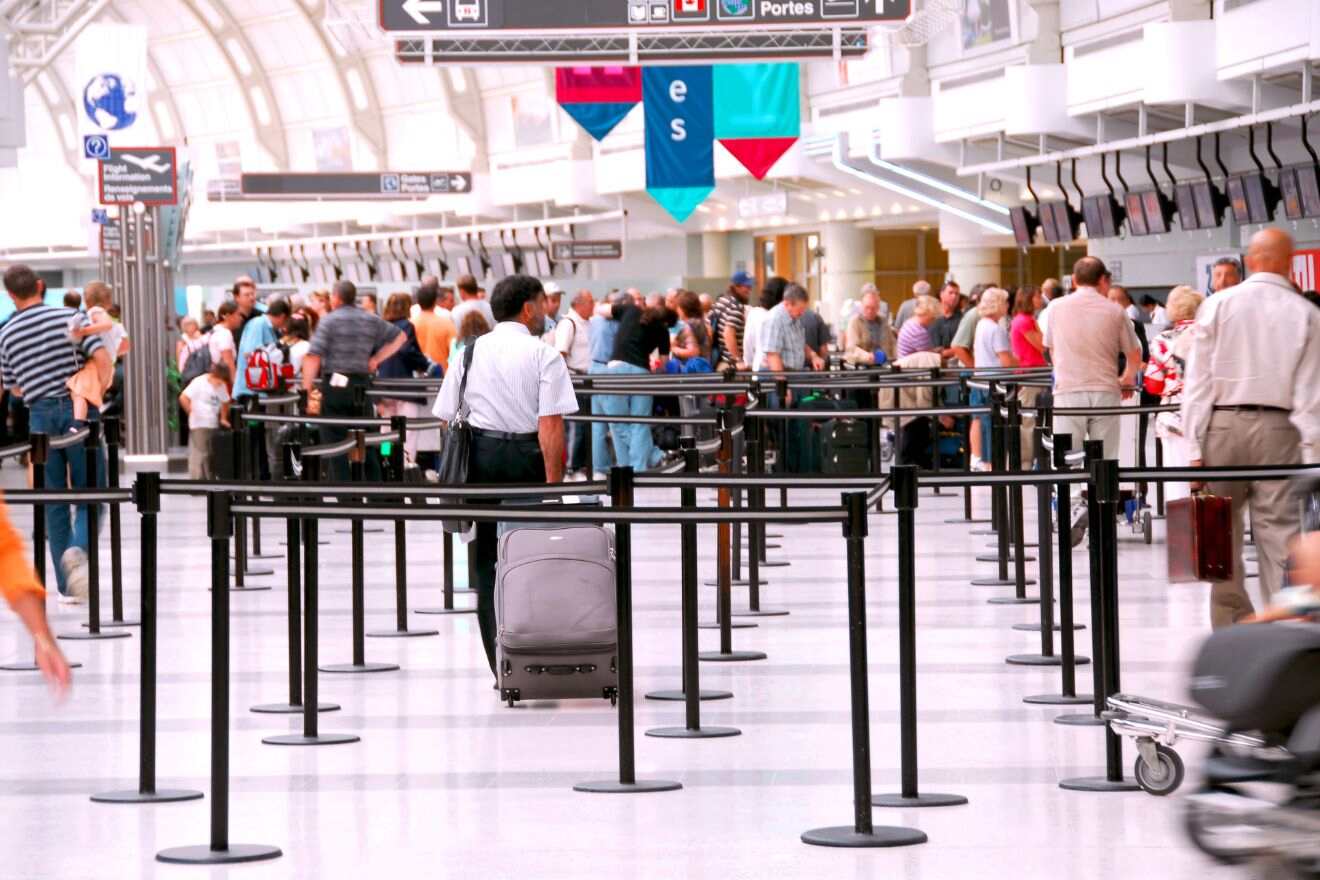 Passengers standing in line at an airport security checkpoint, some pulling suitcases and others carrying bags.