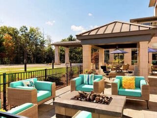 Outdoor patio area with wicker furniture featuring teal cushions, a covered sitting area, a fire pit at the center, and a background view of trees and a road.