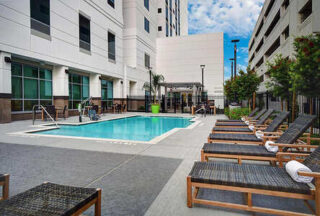 Outdoor hotel pool area with lounge chairs and towels arranged on the deck beside a multi-story building under a partly cloudy sky. Stairs and a lift provide pool access. Some trees are along one side.