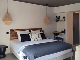 A minimalist bedroom with a double bed, wooden headboard, and monochrome bedding. Pendant lights hang on either side of the bed. A small desk and chair are in the corner.