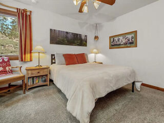 A bedroom with a large bed, two nightstands with lamps, a window with striped curtains, a ceiling fan, and framed pictures on the walls. The room is well-lit with a carpeted floor.