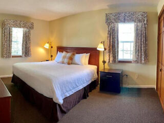 A neatly made bed with white sheets and a plaid pillow is centered between two windows with floral curtains in a small, well-lit bedroom with beige walls and carpeted floor.