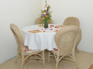 A small round dining table with a white tablecloth, surrounded by wicker chairs and adorned with a vase of colorful flowers in a cozy corner.