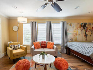 A cozy bedroom with a double bed, a sofa, and a yellow armchair. Two orange poufs and a round coffee table are in front of the sofa. A ceiling fan and three windows with gray curtains are visible.