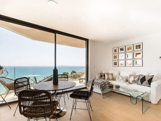 A modern living and dining area with a glass door opens to a balcony with an ocean view. The room has wicker chairs, a round table, a white sofa, a glass coffee table, and framed art on the wall.