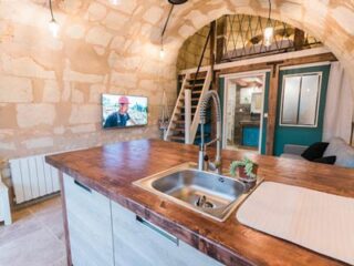 Modern kitchen with a wooden island and stainless-steel sink, stone walls, loft space, and a TV mounted on the wall. The background includes stairs leading to the loft and an entryway.