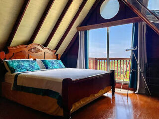 A romantic attic bedroom with wooden beams, a large wooden bed, and a sliding glass door opening to a balcony with a view, featuring a telescope for stargazing.