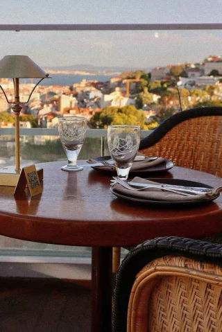 Two empty wine glasses and place settings on a table with a lamp, overlooking a scenic city view from a balcony.