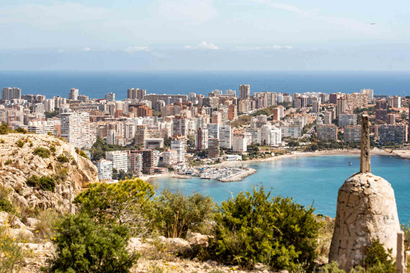 A coastal cityscape with numerous high-rise buildings, a marina, and a rocky hill in the foreground, overlooking a blue sea under a partly cloudy sky.