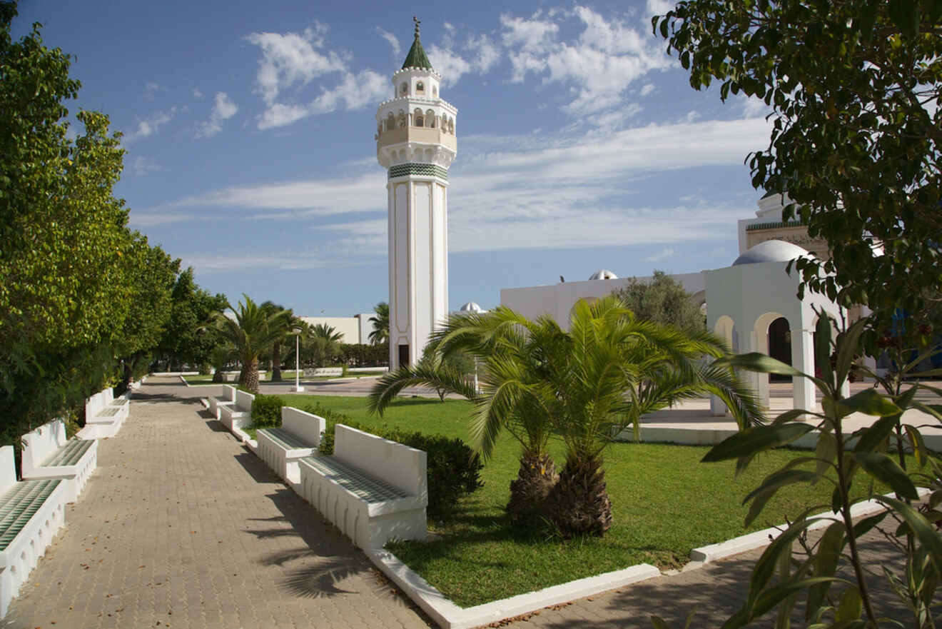 A serene park in Tunis with a white minaret, lush greenery, and a walkway lined with benches under a bright sky.