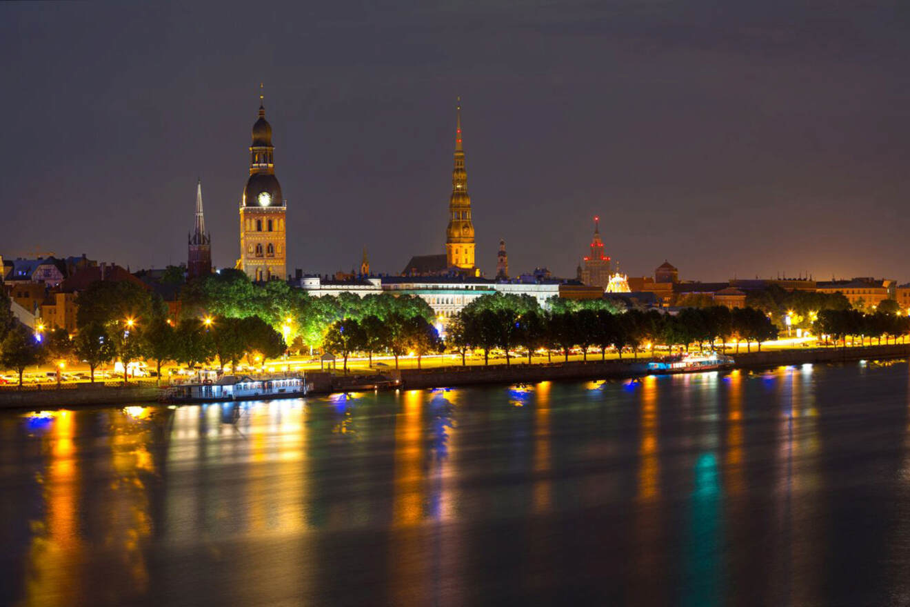 Night view of a cityscape with illuminated buildings, including a cathedral with towers, and their reflections on a calm river.