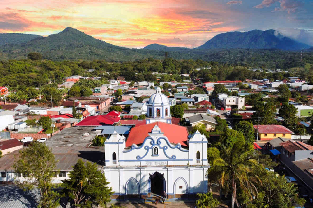 Aerial view of a town with a white church in the foreground, surrounded by buildings and vegetation, with mountains and a vibrant sunset in the background.