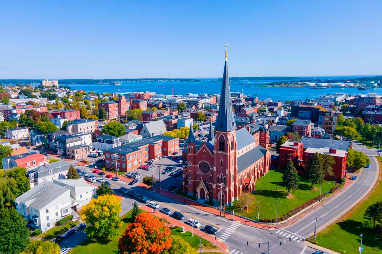 Aerial view of a cityscape featuring a large, red brick church with a tall spire in the center, surrounded by various buildings and roads, with a body of water visible in the distance.