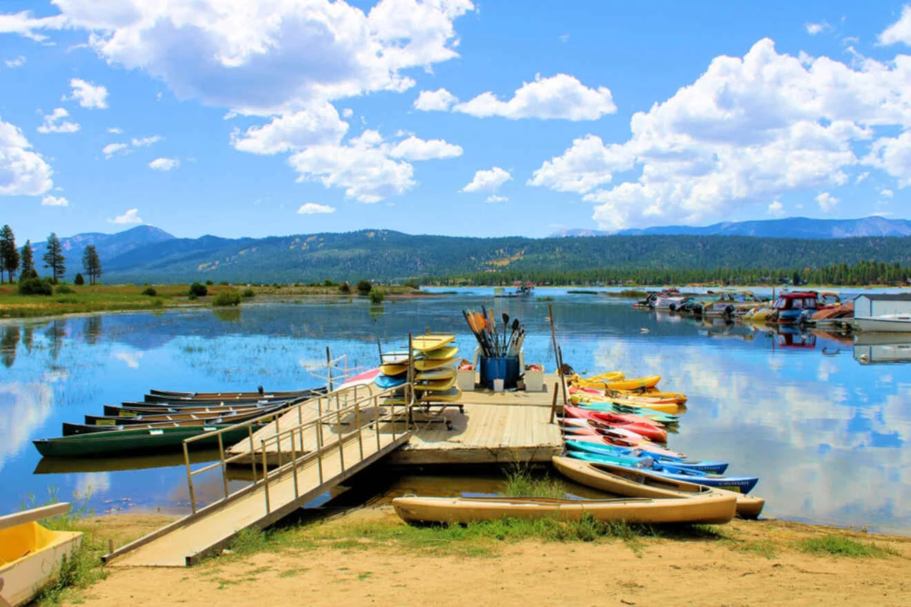 A lake scene with a dock lined with colorful kayaks and canoes, including paddles and other equipment. Mountains and clouds are visible in the background.