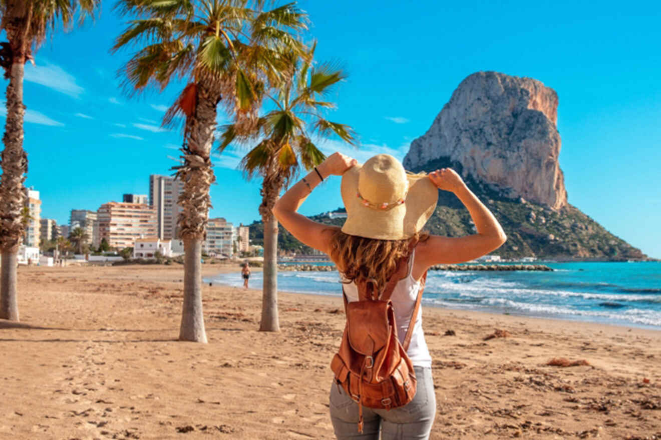 A person wearing a hat and backpack stands on a sandy beach facing the ocean, with palm trees, buildings, and a large rock formation in the background under a clear blue sky.