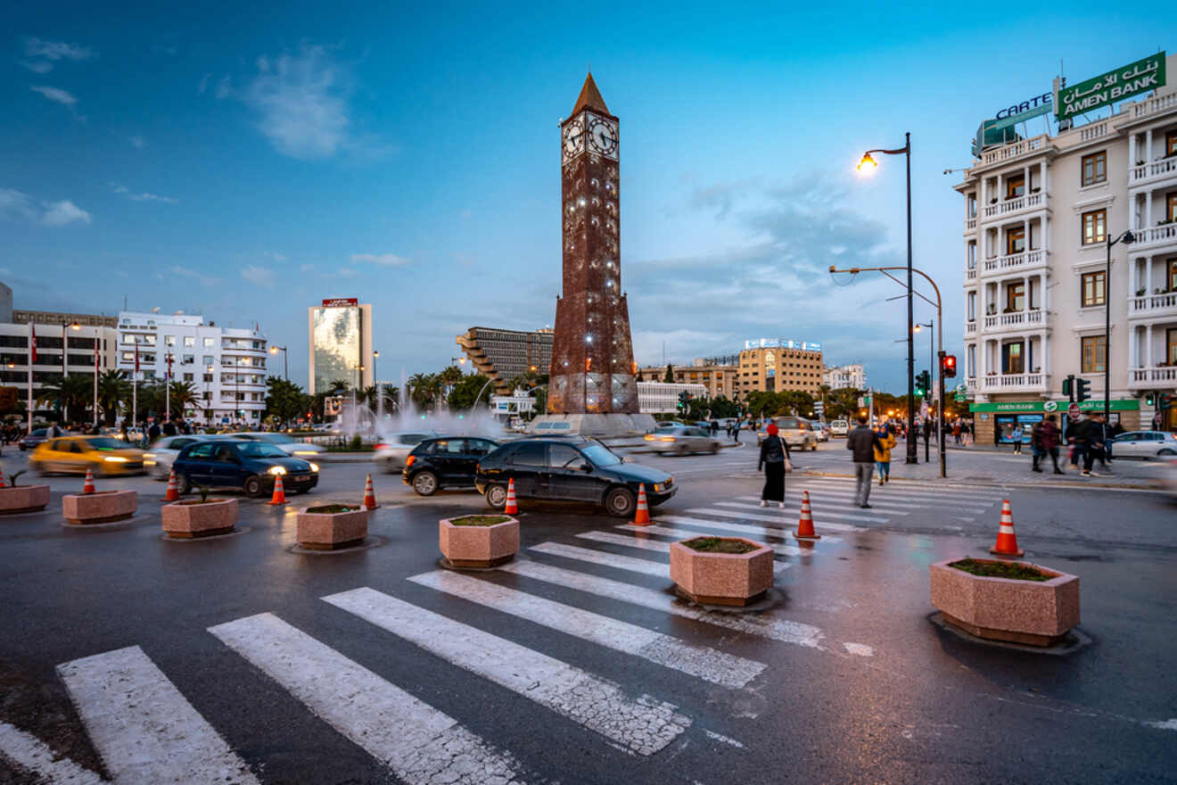 A busy intersection in downtown Tunis with the iconic clock tower, moving traffic, and pedestrians crossing the street at dusk.