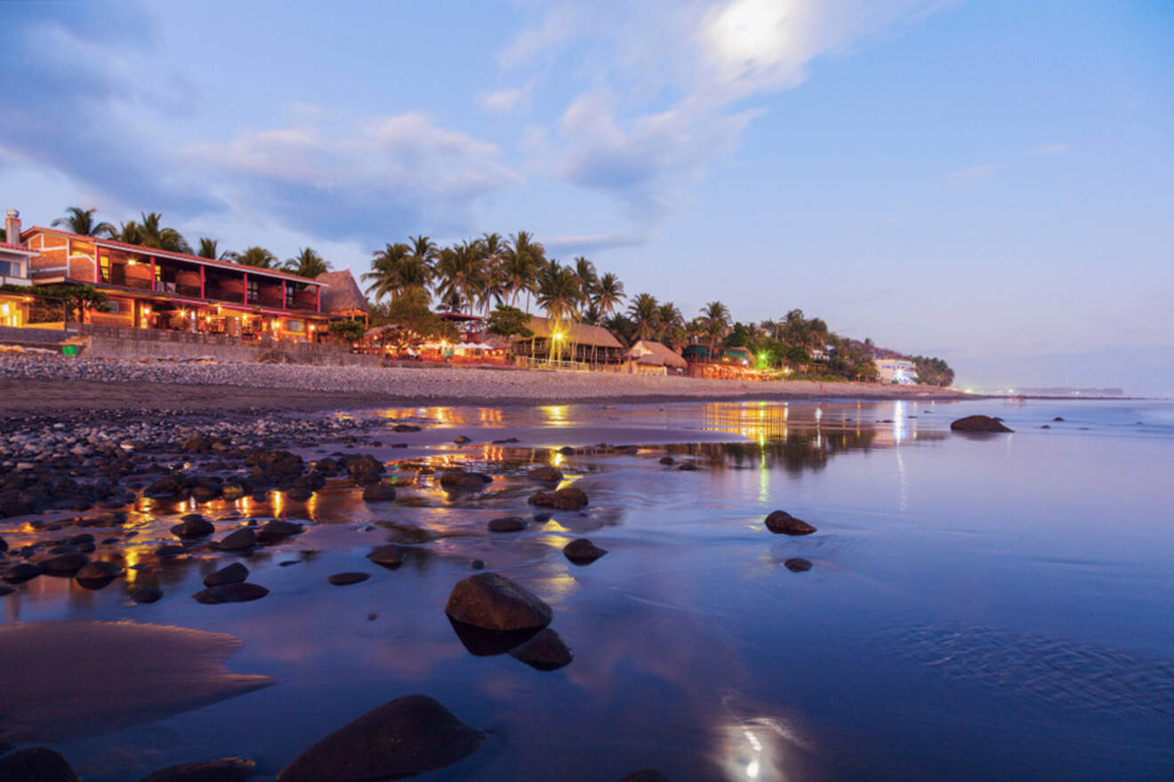 Beach at sunset with calm water, rocky shore, and lit buildings surrounded by palm trees.