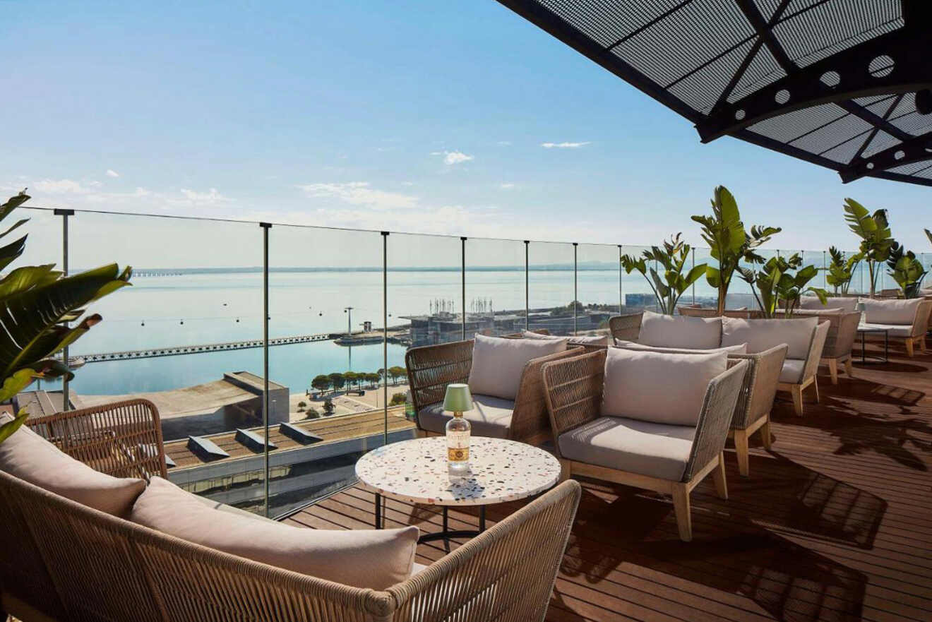 A rooftop lounge area features wicker chairs, a table with a bottle, and potted plants. The space overlooks a clear, expansive body of water under a blue sky.