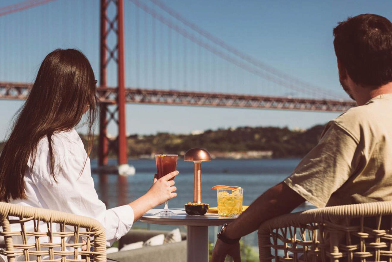 Two people sit at an outdoor table with drinks, overlooking a large red bridge stretching across a body of water under a clear sky.