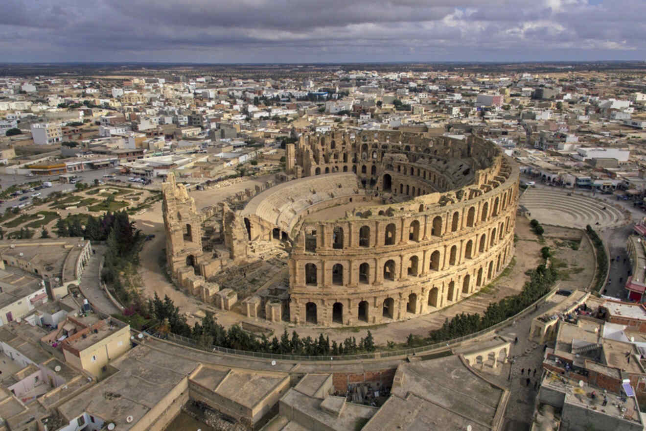 Aerial view of the ancient Roman amphitheater in El Jem, Tunisia, surrounded by the modern cityscape under a cloudy sky.