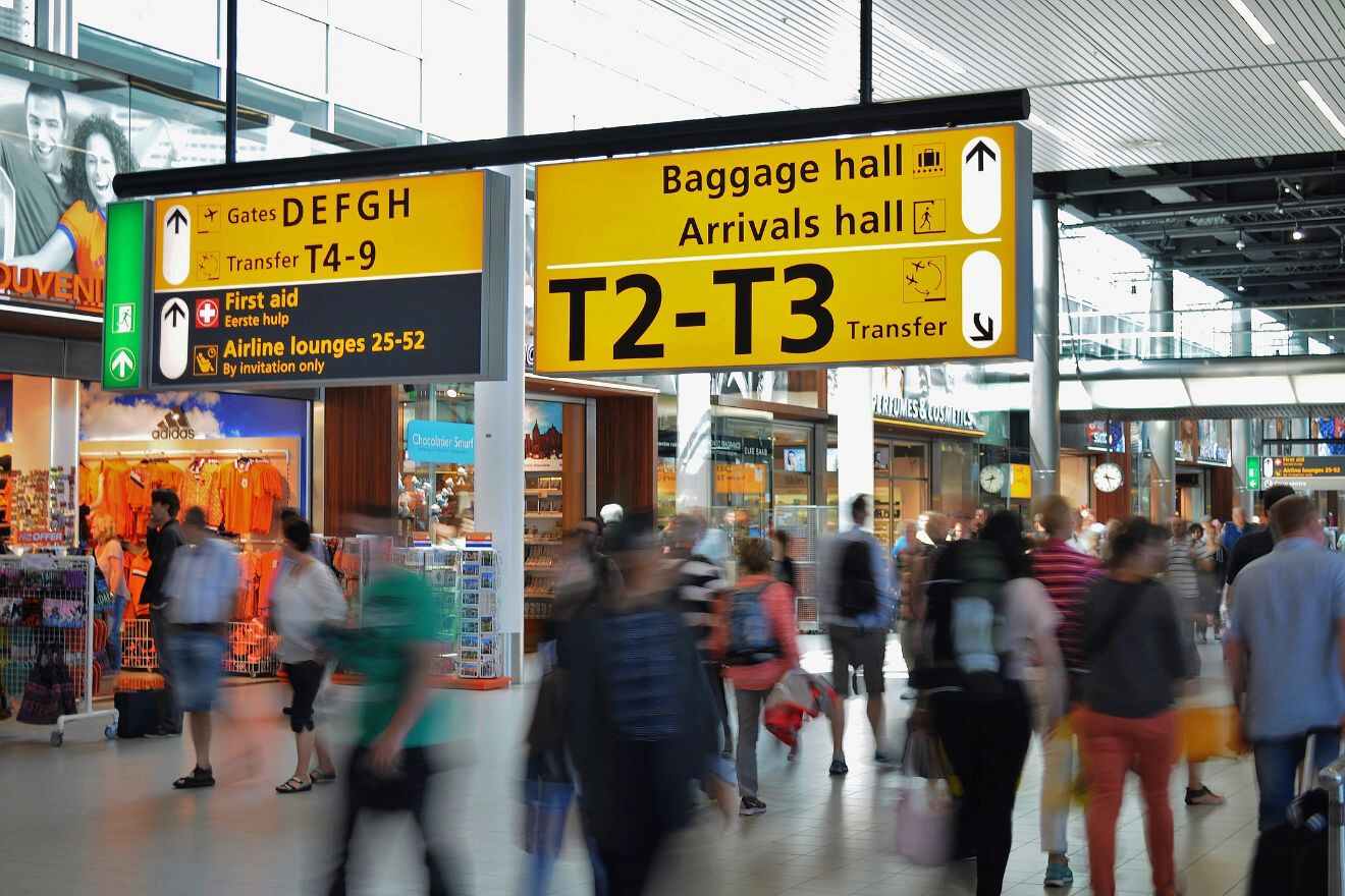 Busy airport terminal with passengers walking, shops in the background, and large overhead signs directing to baggage hall, arrivals, and gates T4-9.