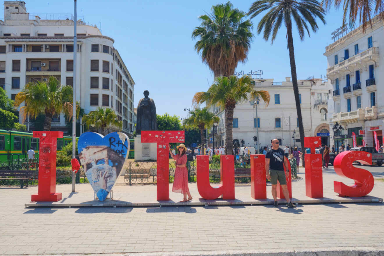 A large "I Love Tunis" sign in a city square with palm trees, people posing, and historic buildings in the background.