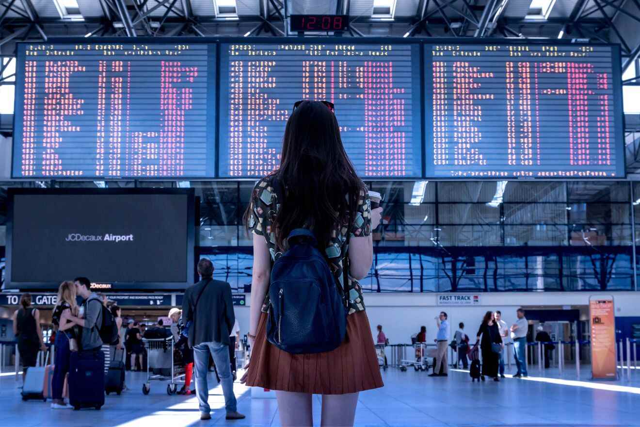 A woman with a backpack stands in an airport terminal, looking at flight information boards displaying various flights and times. Other travelers and airport signs are visible in the background.