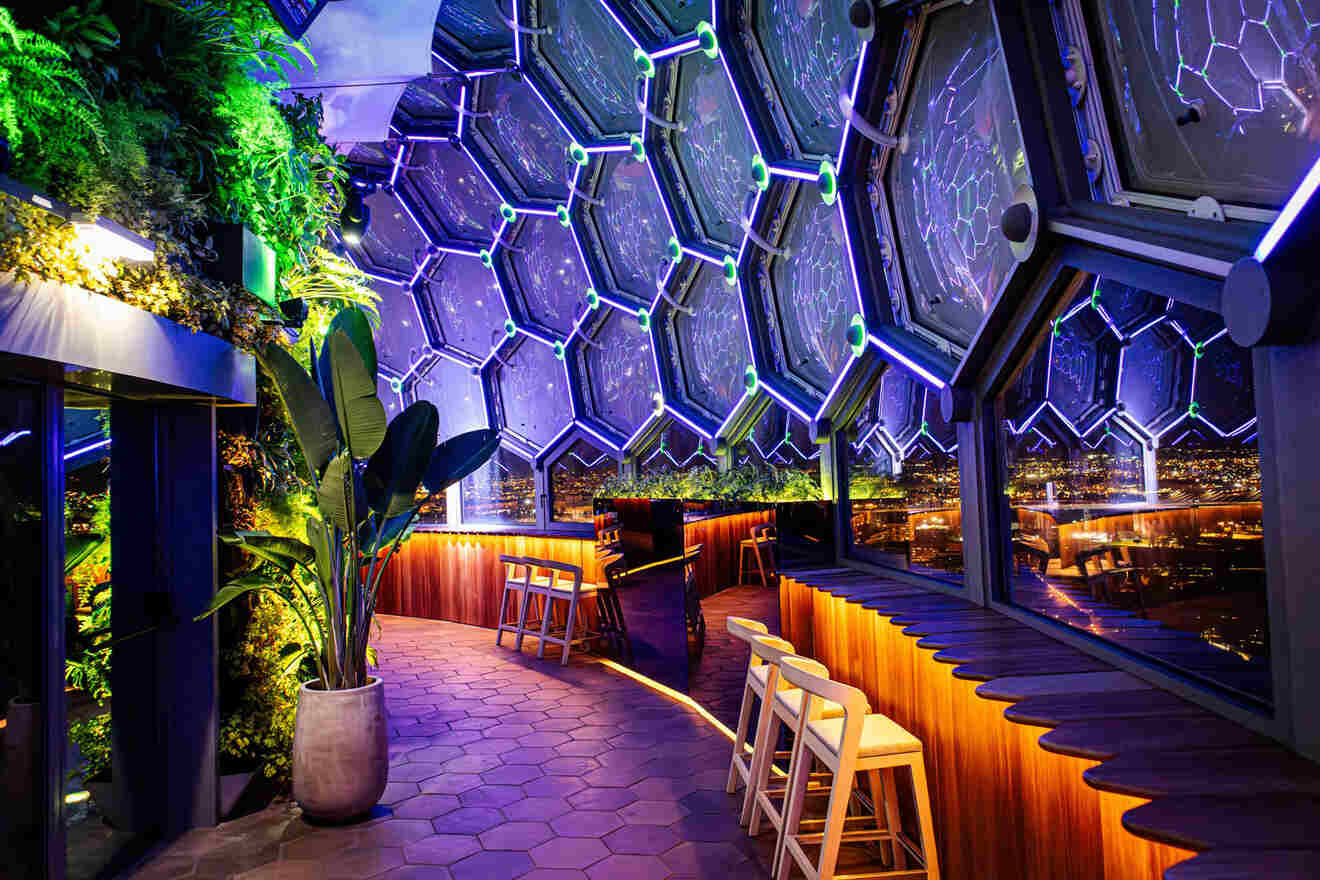 Interior of a modern, hexagon-themed bar with large windows, ambient lighting, a piano, and cityscape views at night.