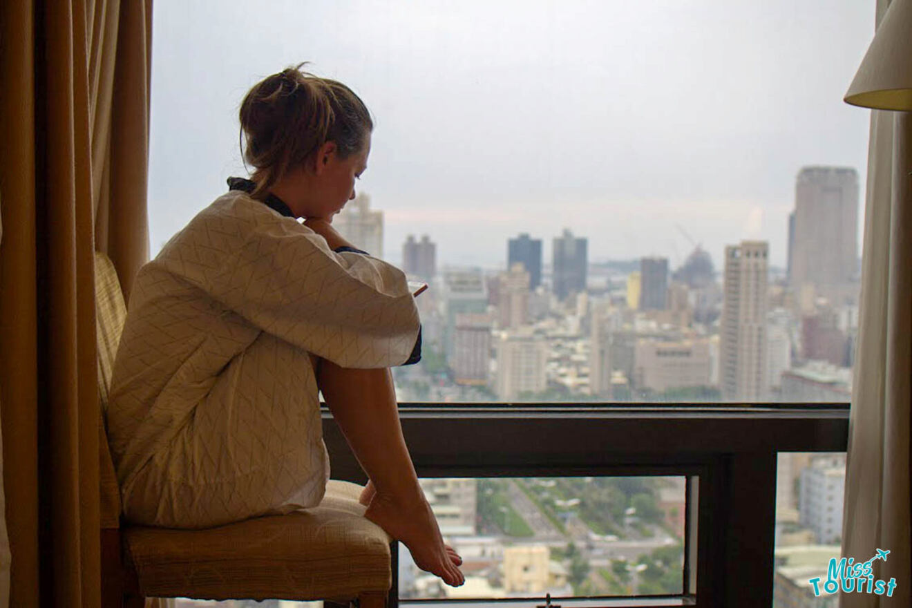 The founder of the page, Yulia, in a robe sits on a chair by a large window, overlooking a cityscape of high-rise buildings.