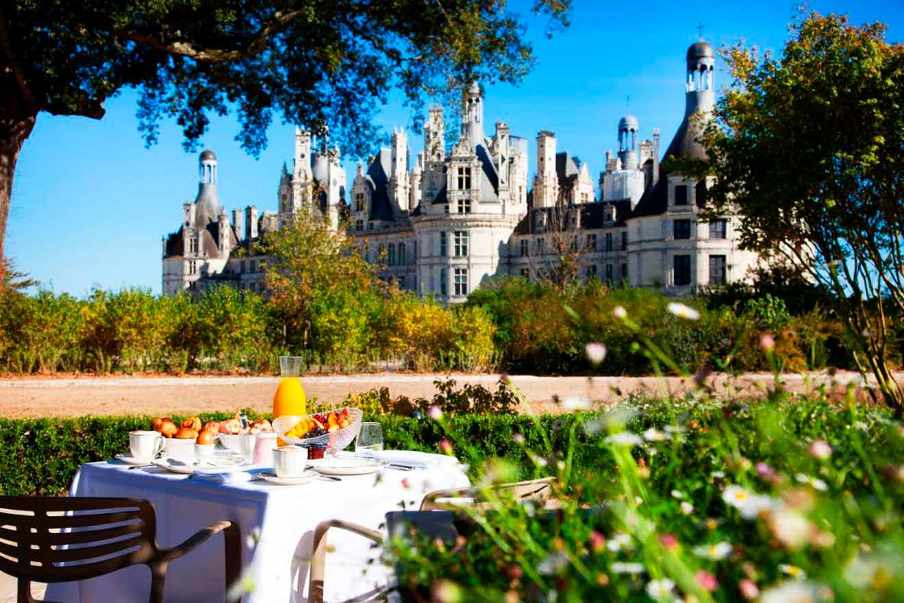 A breakfast table set outdoors in front of a large, historic chateau on a sunny day, surrounded by lush greenery and bright blue skies.