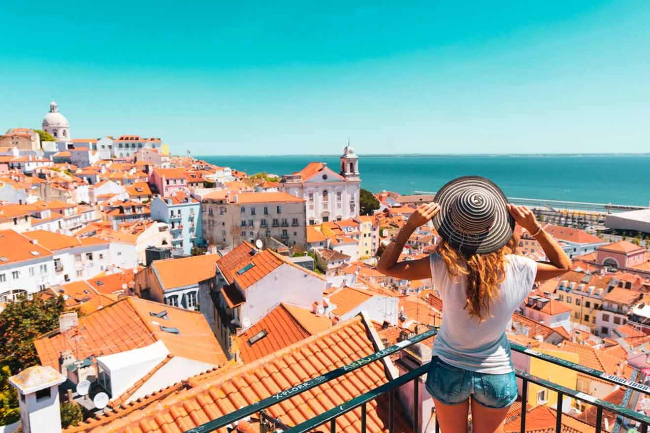 A person wearing a striped hat and shorts looks over a scenic view of a coastal city with red-roofed buildings and the ocean in the background.