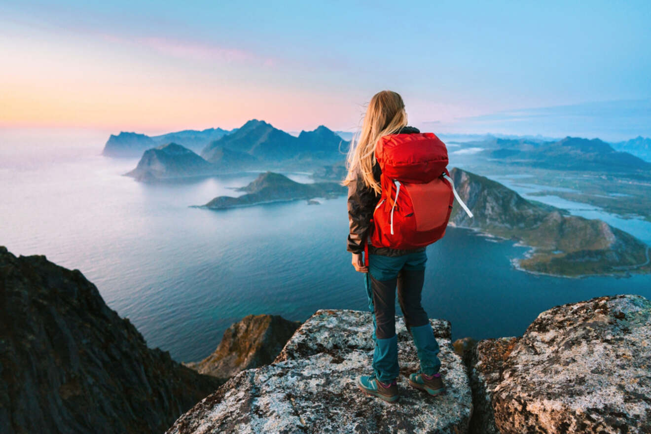 A woman with a red backpack stands on a rocky peak, overlooking a serene coastal landscape with mountains and islands at sunset