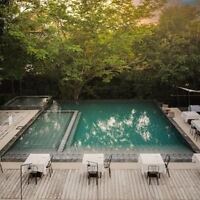 A rectangular outdoor pool surrounded by trees, with adjacent seating areas featuring white tablecloth-covered tables and chairs on a striped patio.