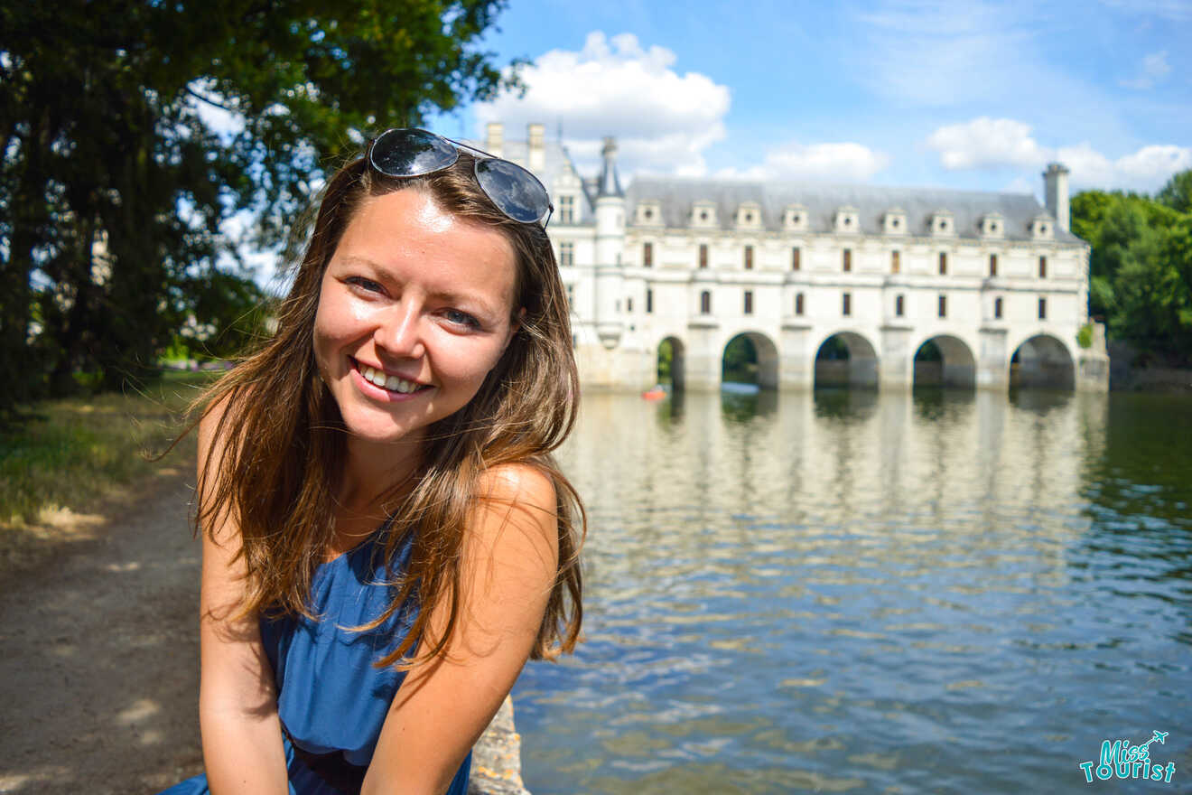 author of the post in blue dress stands in front of Château de Chenonceau, a historic castle over a river, on a sunny day.