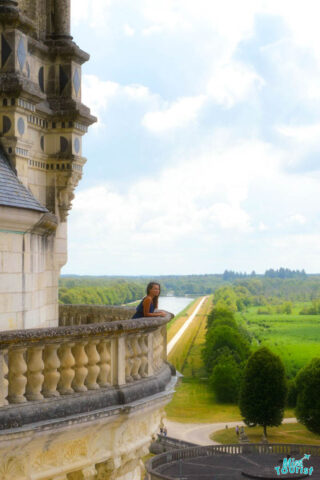 author of the posts stands on a stone balcony overlooking a vast landscape of greenery and a long waterway under a cloudy sky.