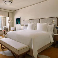 A neatly arranged hotel room with a large bed, white bedding, a cushioned bench at the foot, a modern floor lamp, chairs, a table, and framed artwork on the wall.
