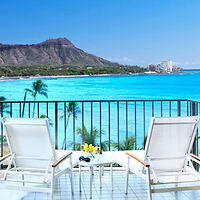 A balcony overlooking a bright blue ocean with two lounge chairs facing the water, a small table with flowers between them, palm trees, and a mountain in the background.