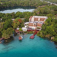 Aerial view of a large building by a lush forest and a body of water with a dock extending into the water and a small boat moored alongside.