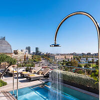 A rooftop pool with an arched shower fixture overlooking a cityscape with buildings under a clear blue sky.