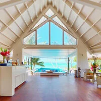 A bright, airy room with high ceilings and large windows showcasing a view of a tropical beach and ocean. The room has modern furnishings, plants, and a bar area on the left.