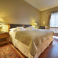 A neatly made double bed with a beige quilt in a warmly-lit bedroom. The room features a wooden floor, bedside tables with lamps, a window with curtains, and a patterned rug.