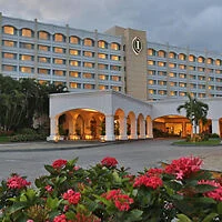 A large hotel building with multiple floors, featuring a prominent entrance and arched drive-through. Red flowers and green foliage are in the foreground.