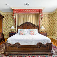 A bedroom with a large wooden bed, white bedding, floral pillows, two side tables with lamps, patterned wallpaper, and a rug on the floor.