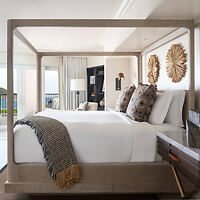 A modern bedroom features a wooden canopy bed with white bedding and patterned pillows. A throw blanket is draped over the end of the bed. The room includes a large window and wall decorations.