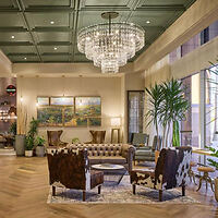 A hotel lobby with a chandelier, green ceiling, leather sofa, and cow-print armchairs. Large windows and plants create a welcoming atmosphere.