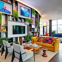 A modern living room with colorful decor, including a yellow sofa, two white armchairs, and vibrant artwork on the walls. The space is well-lit and features various plants and bookshelves.