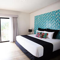 A modern bedroom featuring a king-size bed with white linens, black accents, and colorful pillows. The room has a teal patterned accent wall, a floor lamp, and a sliding glass door with curtains.