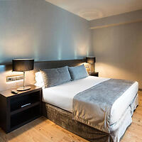 A modern bedroom with a large bed, two bedside tables with lamps, and gray bedding against a plain wall.