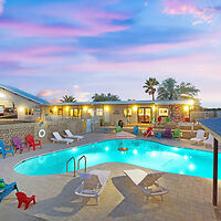 A well-lit outdoor pool area with lounge chairs and colorful seating surrounds a clear blue pool. A building with lit windows is visible in the background, all under a twilight sky.